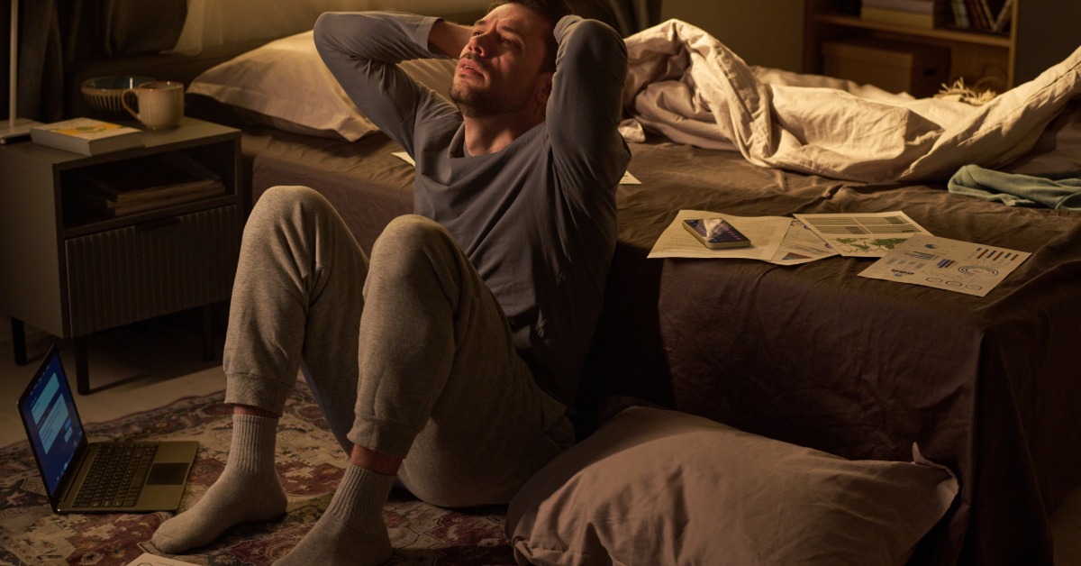 A man sits on the floor by an unmade bed, his hands behind his head. A laptop and scattered papers are on the floor.