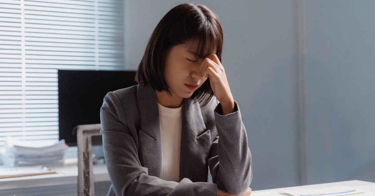 A woman sits in an office with her eyes closed, pressing the bridge of her nose. Documents are spread out on her desk.