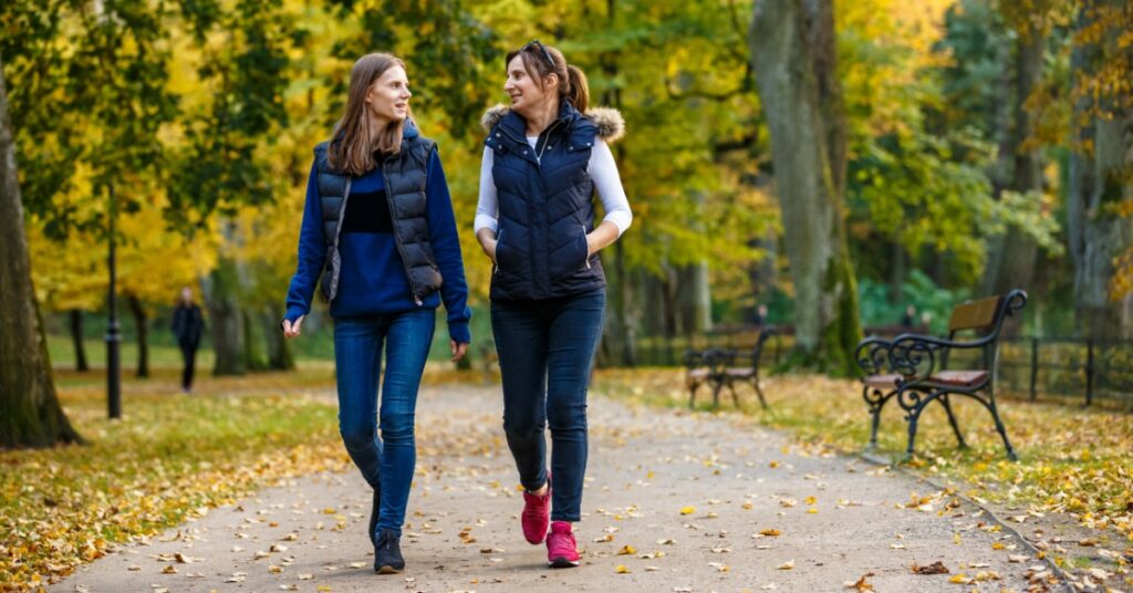 Two women walk together on a leaf-covered park path. Benches and trees with green and yellow leaves line the walkway.