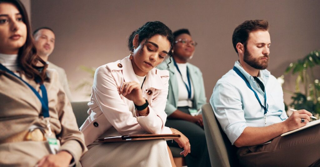 A group of adults sits at an indoor event or meeting. Most of them watch the presenter, but one person checks her watch.