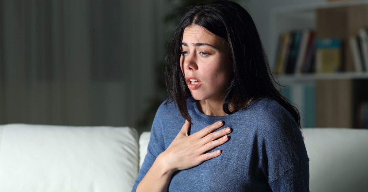 A woman sits upright on a couch and presses one hand to her chest. Her mouth is slightly open as she looks downward.