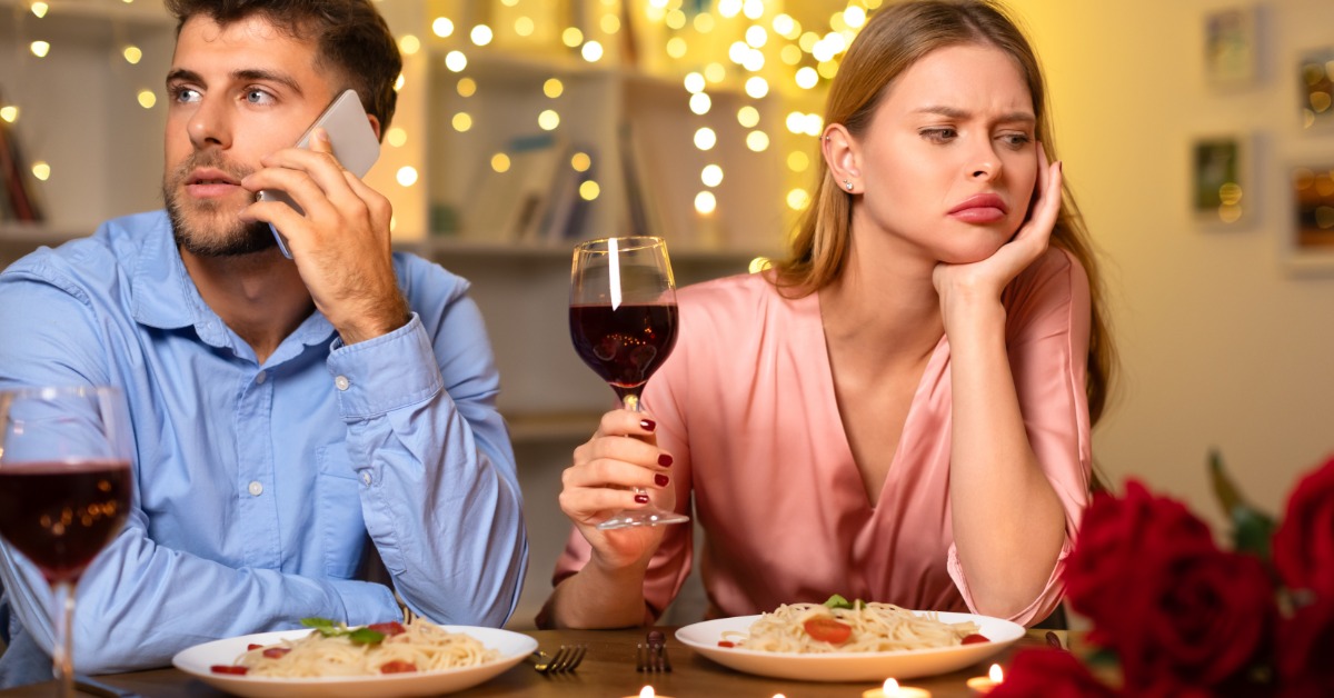 A man and a woman at a dinner table face away from each other. The man talks on his cell phone, and the woman frowns.