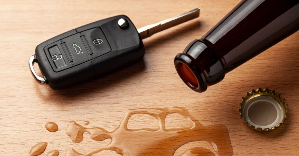 A dark glass bottle lies on its side next to car keys. Spilled liquid under the bottle forms the shape of a crashed car.