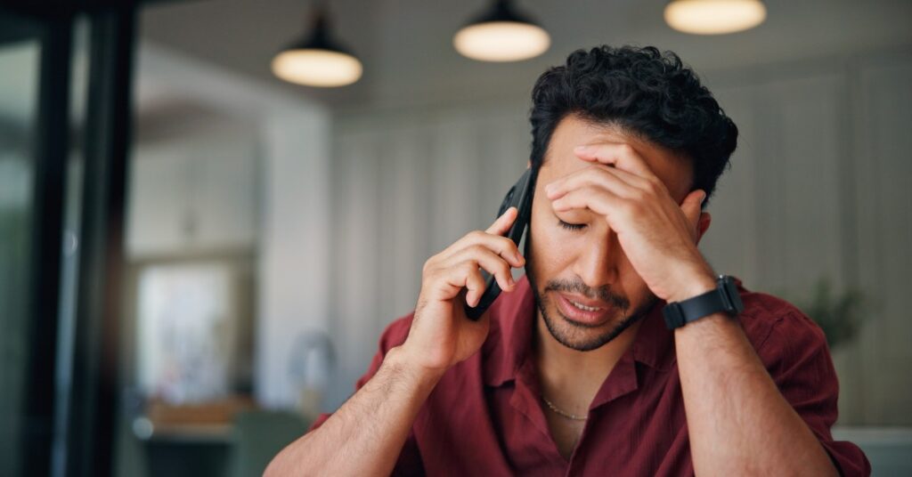 A seated man holds a smartphone to his ear and presses his other hand against his forehead. His eyes are closed.