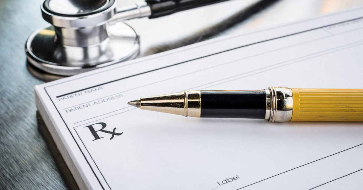 A black-and-yellow pen rests across a prescription form marked with an “Rx” symbol. A stethoscope lies above the form.