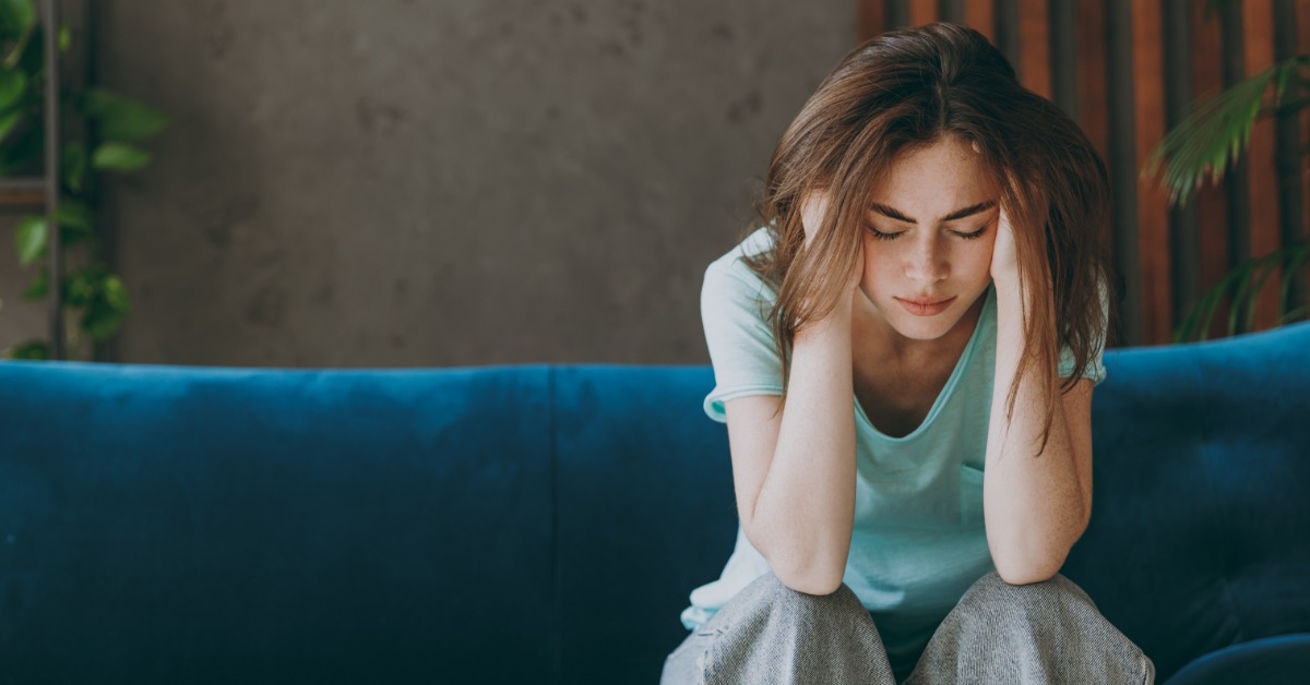 A woman on a couch leans forward, elbows on her knees, eyes closed. She pushes both hands against the sides of her head.