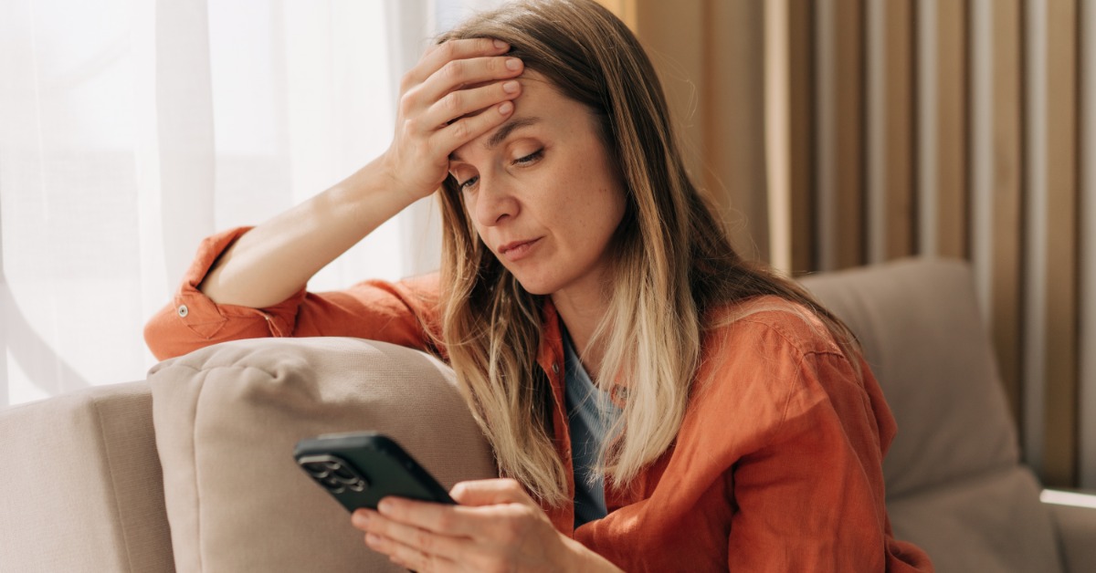 A woman sits on a couch beside a window. She looks at a phone she holds and presses her other palm to her forehead.