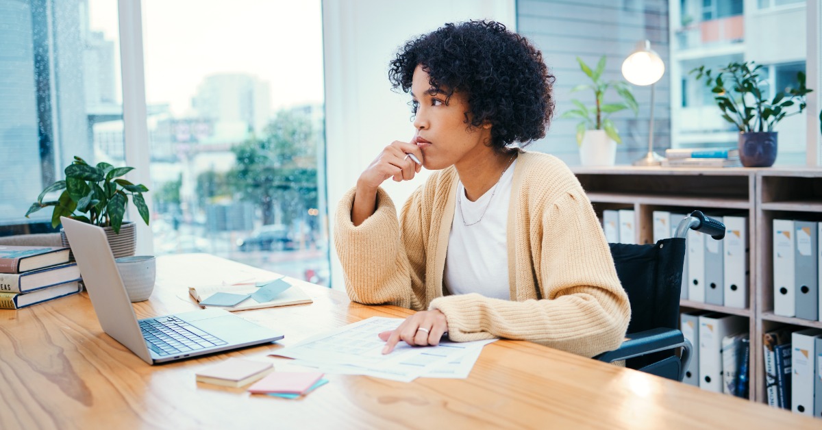 A woman sits at a desk with notepads, an open laptop, and papers. Her index finger is on one page, and she looks away.
