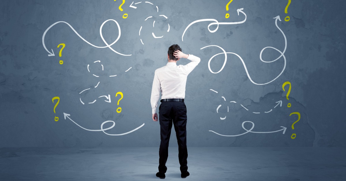 A man faces a blue-gray wall with one hand on the back of his head. Several question marks and arrows are on the wall.