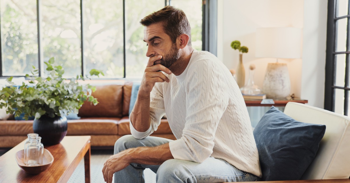 A man in a white sweater leans slightly forward on a couch. He rests one hand against his mouth and looks down.