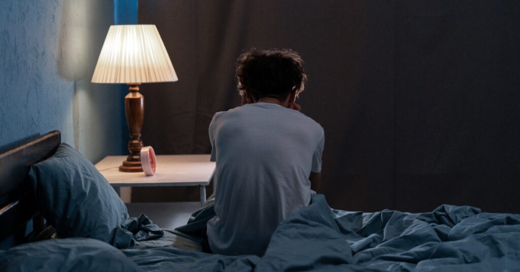 A person sits on the edge of a bed with their back to the camera. A lit bedside lamp is on the nightstand beside them.
