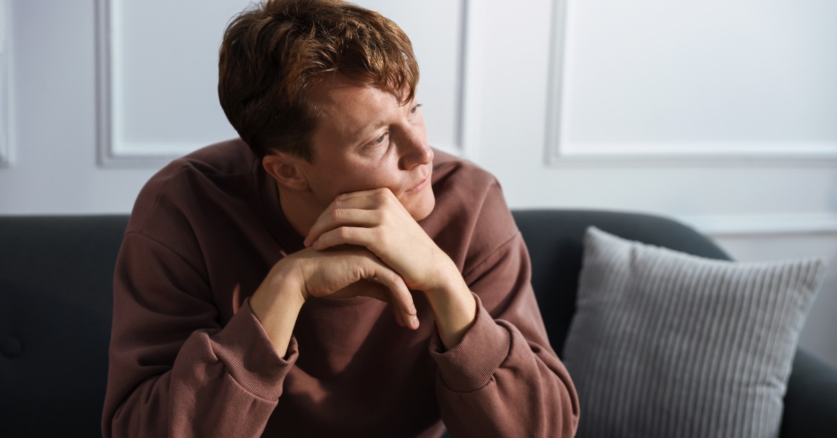 A young man in a brown sweatshirt leans forward on a dark sofa, chin resting on clasped hands. He looks off to the side.