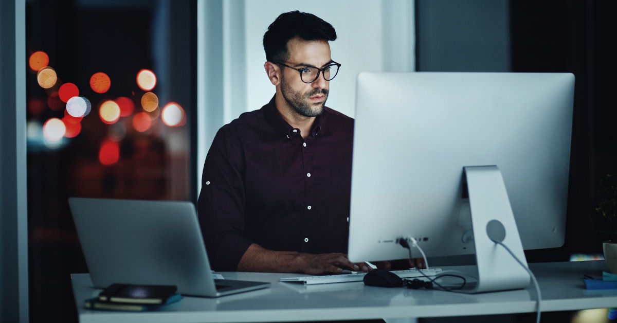 A man at a desk looks at a large computer monitor. Blurred city lights are visible through the window behind him.