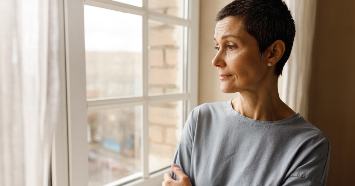 A woman stands near a white-framed window and gazes out. Her arms are folded loosely across her chest.